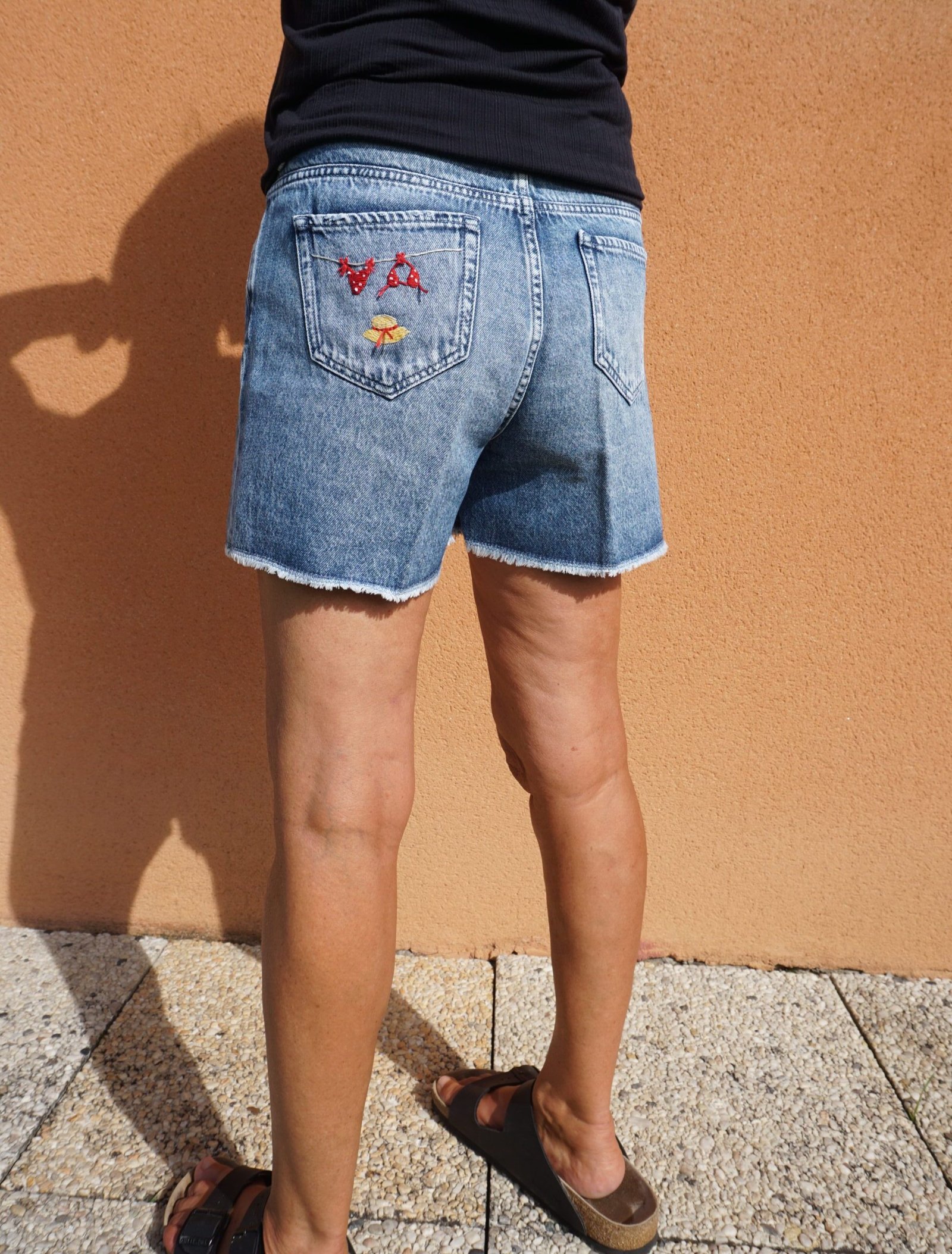 broderie à la main d'un bikini rouge à pois et d'un chapeau de paille sur la poche arrière d'un short en jean porté, vu de profil