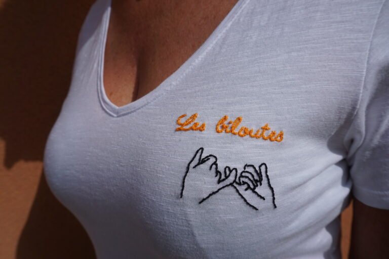 gros plan sur la broderie à la main de l'inscription Les biloutes en fil orange et de deux mains liées par le petit doigt sur un tee-shirt blanc
