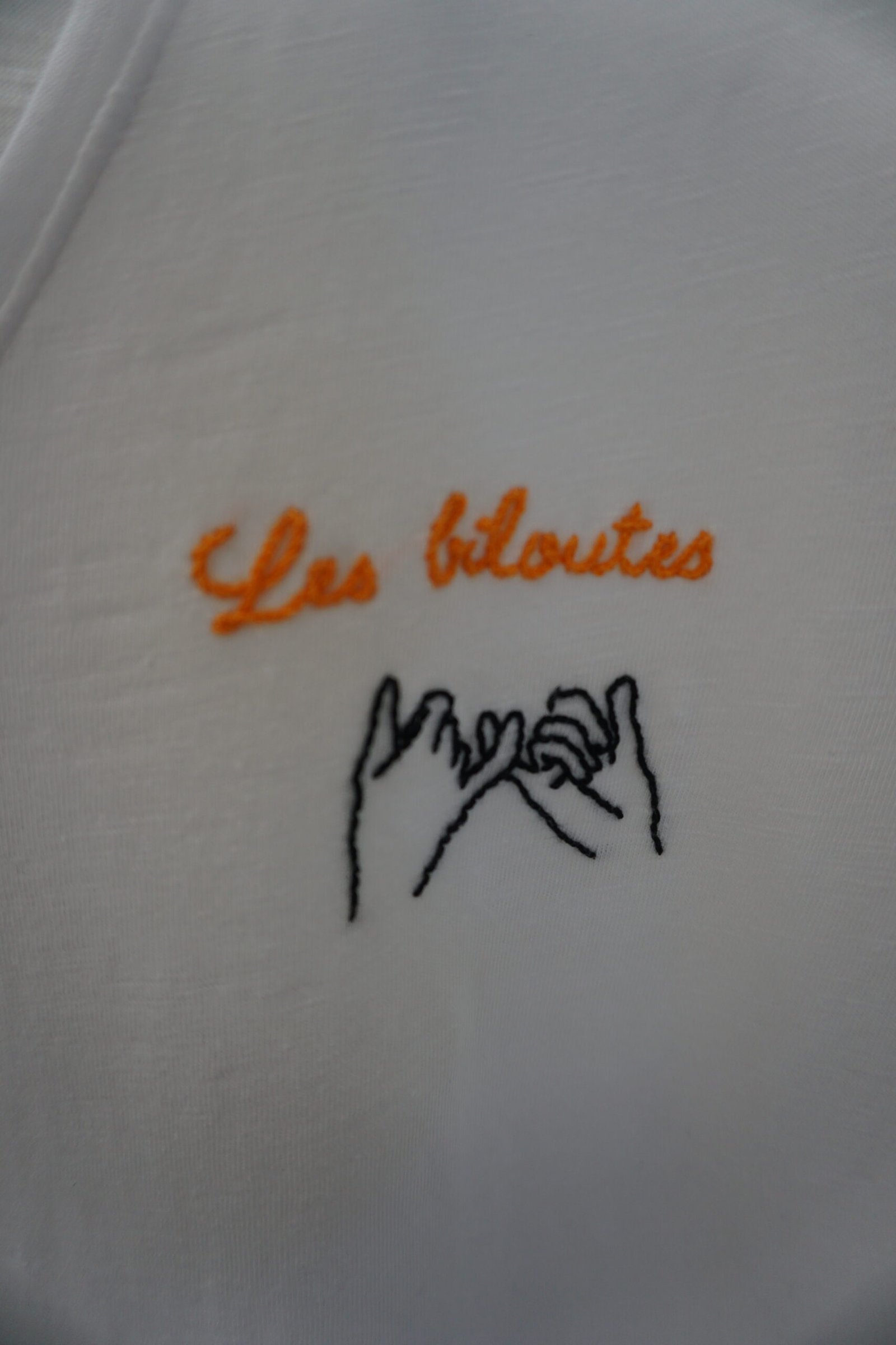 broderie à la main de l'inscription Les biloutes en fil orange et de deux mains liées par le petit doigt sur un tee-shirt blanc à plat